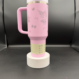 Puede incluir: Taza de viaje aislada rosa con asa, con un dise&ntilde;o de personaje de dibujos animados y detalles de lazo. La taza tiene una tapa blanca y una banda plateada. Est&aacute; sobre una base circular blanca.