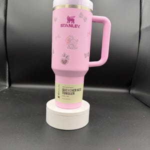 Puede incluir: Un vaso Stanley Quencher H2.0 rosa con asa y pajita. El vaso presenta el logotipo de Stanley e ilustraciones decorativas. El texto "THE FLOWSTATE QUENCHER H2.0 TUMBLER" est&aacute; impreso en una etiqueta cerca de la base. El vaso se muestra sobre un soporte blanco.