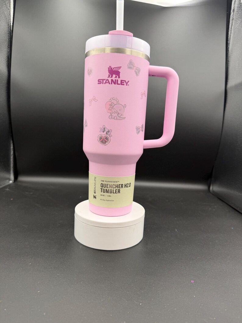 Puede incluir: Un vaso Stanley Quencher H2.0 rosa con asa y pajita. El vaso presenta el logotipo de Stanley e ilustraciones decorativas. El texto "THE FLOWSTATE QUENCHER H2.0 TUMBLER" est&aacute; impreso en una etiqueta cerca de la base. El vaso se muestra sobre un soporte blanco.