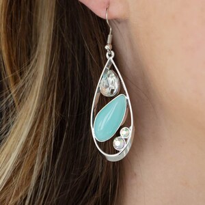 May include: Silver-toned teardrop earrings with a clear, teardrop-shaped gem at the top, a light blue oval gem in the center, and two small, iridescent gems at the bottom. The earrings are worn on a person's ear.