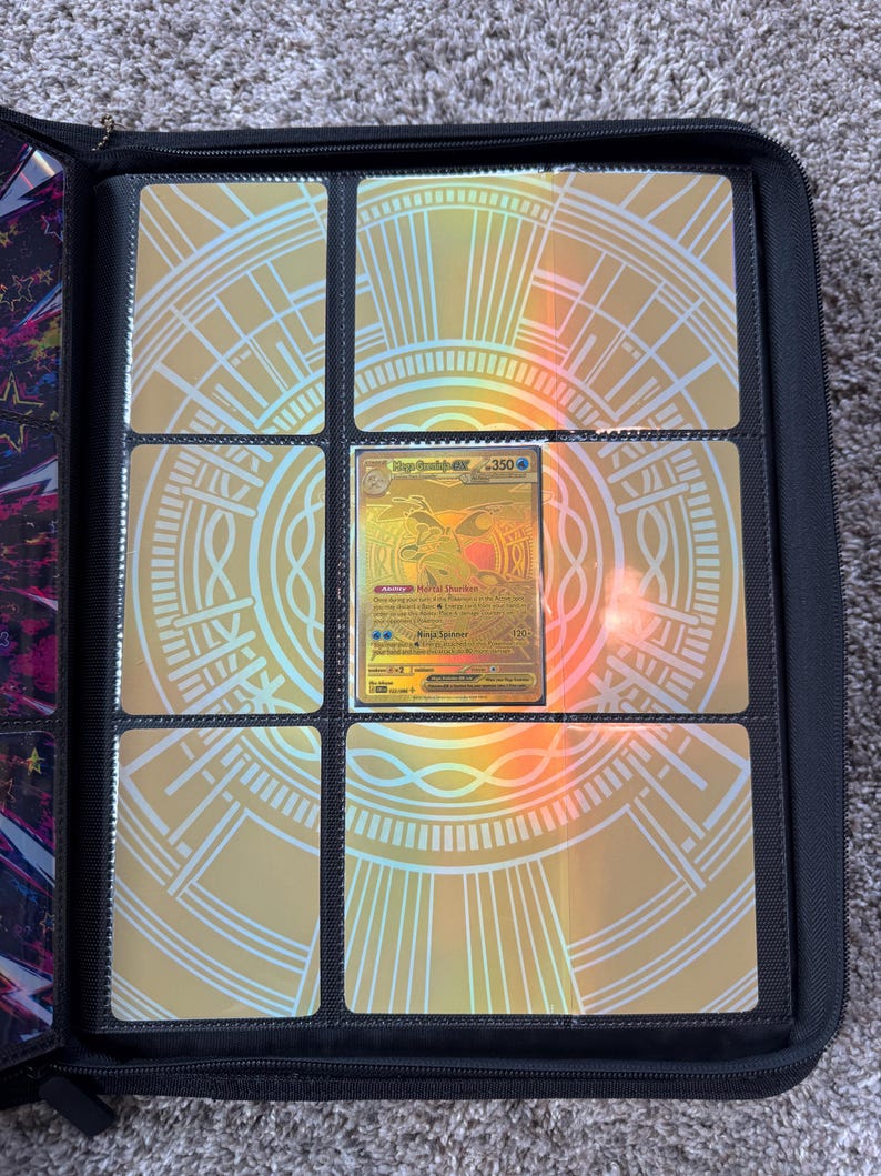 May include: A black zippered trading card binder featuring a gold holographic insert with a circular geometric pattern. A single gold-colored Mega Greninja GX trading card with 350 HP is displayed in the center pocket of the nine-pocket page.