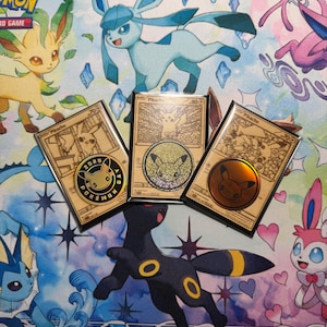 May include: Three gold-colored Pikachu trading cards in protective sleeves, each featuring a unique circular metallic coin emblem. The cards display intricate line art of Pikachu and are arranged on a colorful playmat featuring various Pokémon characters.