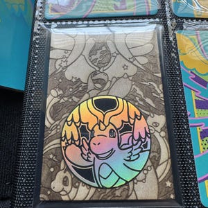 Könnte beinhalten: Eine rechteckige Sammelkarte mit einem hellbraunen Holzmaserungshintergrund und einem sich wiederholenden Muster aus drachenähnlichen Comicfiguren und Sternen. Ein kreisförmiger, schillernder, regenbogenfarbener Aufkleber eines geflügelten Drachen ist mittig platziert.