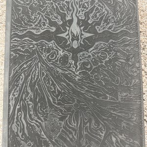 May include: A dark gray rectangular card binder featuring an embossed, intricate fantasy-style design of a central starburst, swirling patterns, and a small knight helmet emblem at the bottom. The text CARD GUARDIAN is embossed at the bottom.
