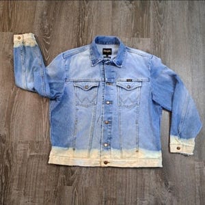 May include: Light blue denim jacket with a faded, bleached hem and cuffs. The jacket features two chest pockets, two side pockets, and a button-up front. The Wrangler logo is visible on the collar.