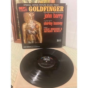 May include: A vintage vinyl record and album cover for the "Goldfinger" soundtrack. The album cover features a woman with gold body paint and the text "Goldfinger" and "John Barry". The record is black.
