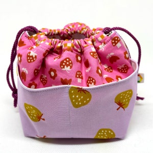 May include: A small drawstring bag. The exterior is lavender, patterned with large, yellow-green strawberries. The interior is pink, with a pattern of mushrooms, flowers, and strawberries. The drawstring is a dark purple. The bag is approximately 10 cm high.