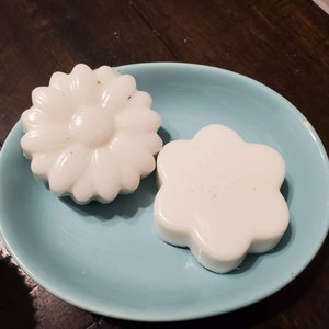 May include: Two white soap bars shaped like flowers on a light blue plate. The soap bars are on a wooden surface.