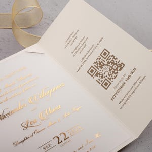 Könnte beinhalten: Eine offene Hochzeitseinladung mit goldfarbenen Buchstaben und einem QR-Code. Die Einladung enthält die Namen des Paares, das Datum und den Veranstaltungsort. Im Hintergrund ist ein goldenes Band zu sehen.