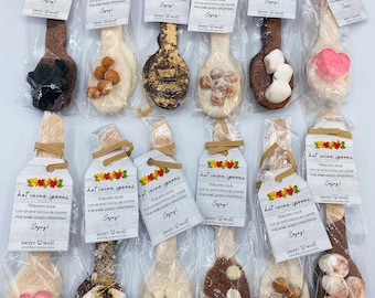 Op de afbeelding: Een assortiment warme chocoladelepels, elk afzonderlijk verpakt en versierd met verschillende toppings zoals marshmallows, chocoladeschilfers en noten. Elke lepel heeft een label met de tekst "hot cocoa spoons" en "Enjoy!"