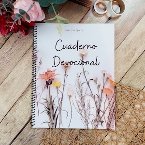 May include: A spiral-bound notebook with the words "Cuaderno Devocional" and floral illustrations. The notebook is surrounded by colored pencils, flowers, and decorative rings. The background is a wooden surface.