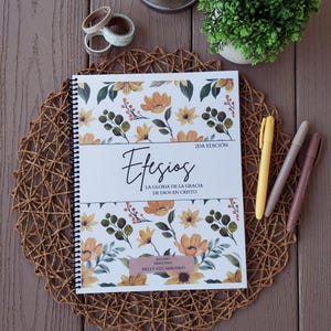 May include: A spiral-bound notebook with a floral design featuring orange and yellow flowers and green leaves. The cover has the title "Efesios" and other text in Spanish. A yellow pen, a gray pen, and a pink pen are next to the notebook.