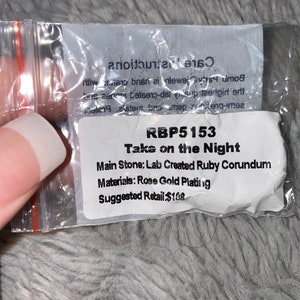 May include: A white card with the text "RBP5153 Take on the Night" and details about the jewellery piece. The main stone is a lab-created ruby corundum, the materials are rose gold plating, and the suggested retail price is &pound;108.