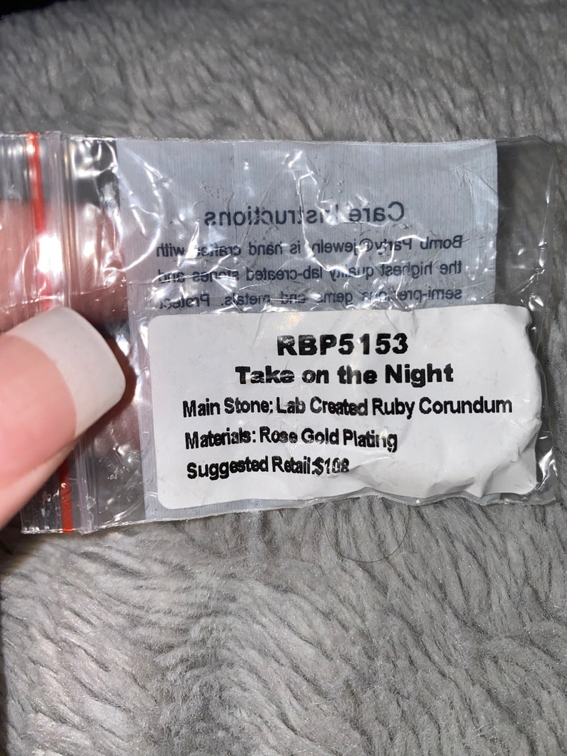 May include: A white card with the text "RBP5153 Take on the Night" and details about the jewellery piece. The main stone is a lab-created ruby corundum, the materials are rose gold plating, and the suggested retail price is &pound;108.