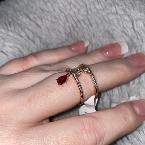 May include: A gold ring with a teardrop-shaped red stone dangling from it. The ring is set with small clear stones and has a delicate design.