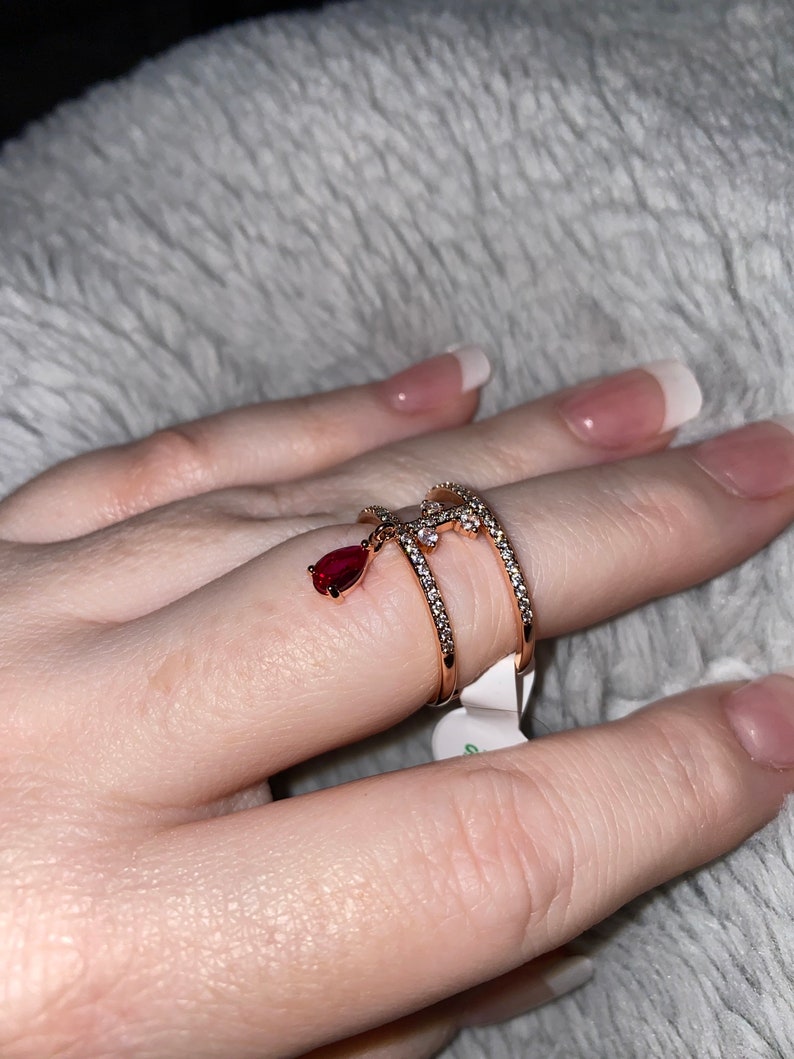 May include: A gold ring with a teardrop-shaped red stone dangling from it. The ring is set with small clear stones and has a delicate design.