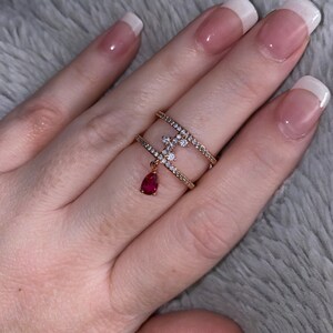 May include: A gold ring with a red stone pendant and clear stones. The ring is double-banded and has a delicate design.