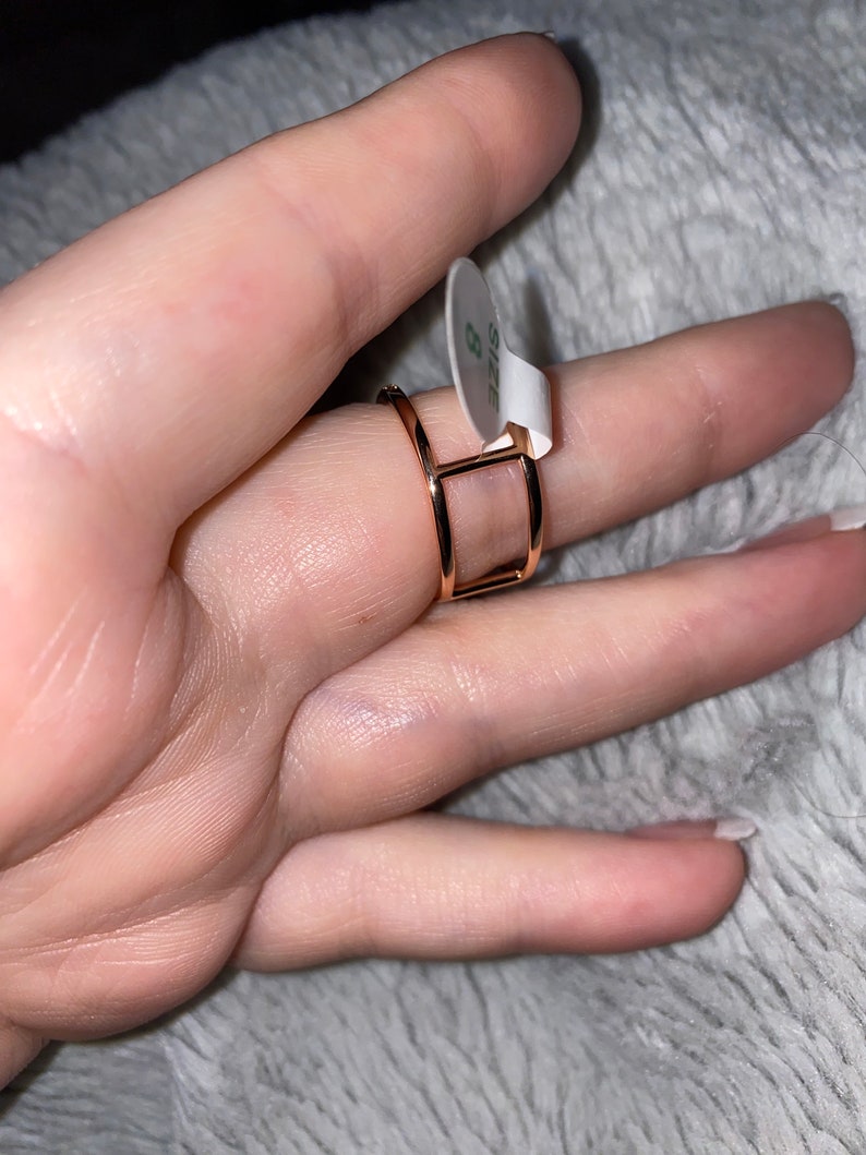 May include: A gold-toned ring with a square design. The ring is on a finger and has a size 8 tag attached.