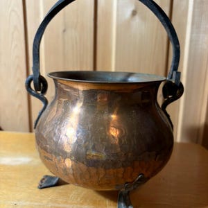 Può includere: Un calderone in rame martellato con un manico in ferro nero e tre piedini decorativi. Il calderone ha un corpo arrotondato e una patina scura. Il manico è arcuato e fissato con anelli decorativi. Stile vintage.