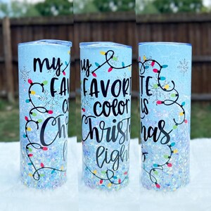 May include: A blue tumbler with glitter and a white snowflake design. The tumbler has the text "My favorite color is Christmas lights" in black.