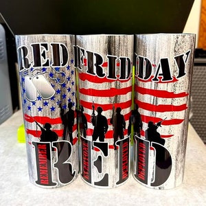 May include: Three stainless steel tumblers with a patriotic design. The tumblers feature the words "RED FRIDAY" in bold black letters. One tumbler has a blue star pattern, and the others have a red and white stripe design. Silhouettes of soldiers are also visible.