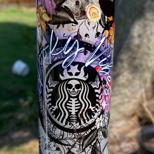 May include: A tall, stainless steel tumbler with a Halloween-themed design. The design includes a skeleton in the Starbucks logo, purple flowers, spiderwebs, and the words "Spooky Vibes". The tumbler is approximately 9 inches tall.