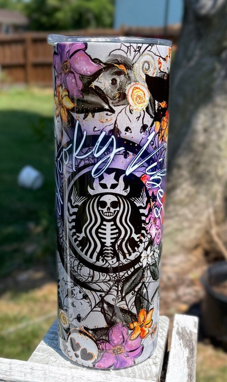 May include: A tall, stainless steel tumbler with a Halloween-themed design. The design includes a skeleton in the Starbucks logo, purple flowers, spiderwebs, and the words "Spooky Vibes". The tumbler is approximately 9 inches tall.