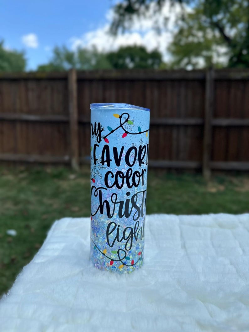 May include: A blue tumbler with glitter and a white design that says "My favorite color Christmas lights".