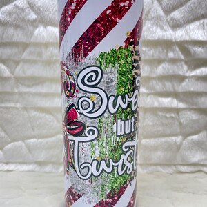 May include: A stainless steel tumbler with a festive design. The tumbler features red glitter stripes, a bow, and the words "Sweet but Twisted" in a whimsical font. The design includes green glitter and a leopard print.