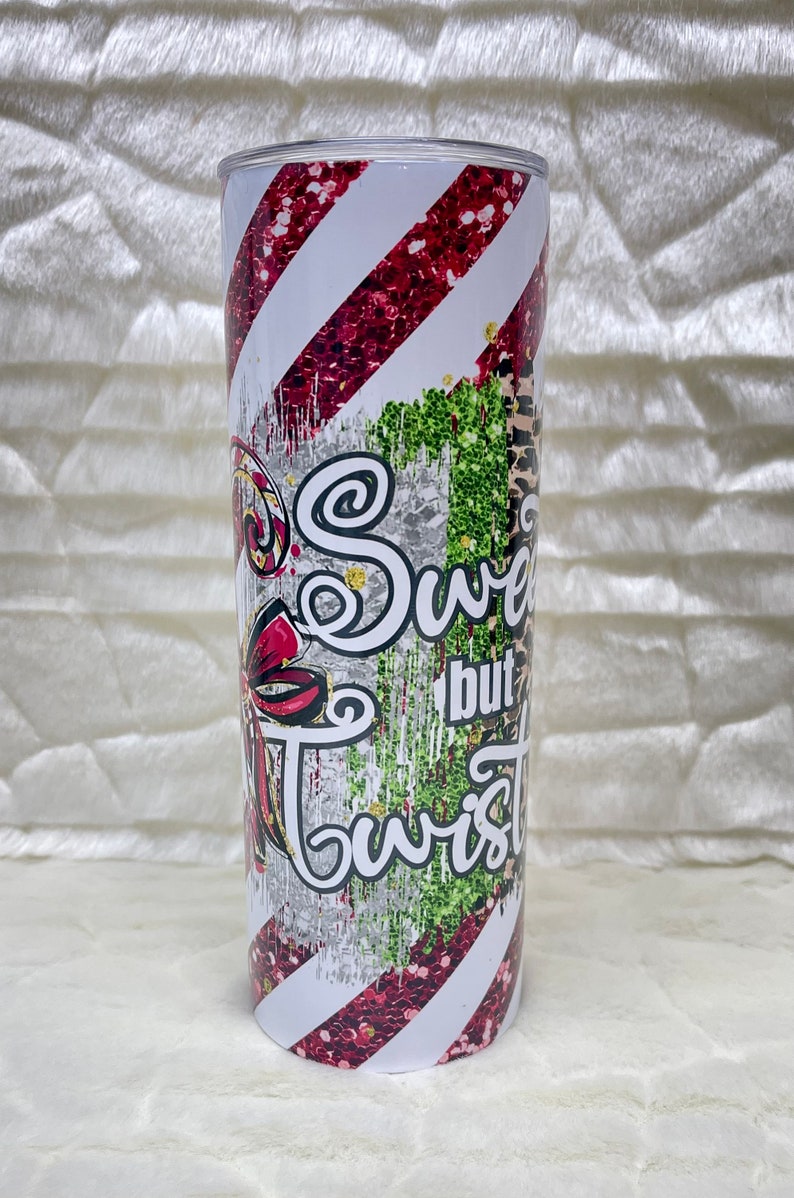 May include: A stainless steel tumbler with a festive design. The tumbler features red glitter stripes, a bow, and the words "Sweet but Twisted" in a whimsical font. The design includes green glitter and a leopard print.