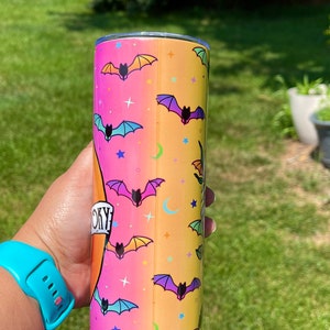 May include: A Halloween-themed tumbler with a gradient background of pink, orange, and yellow. The tumbler is decorated with colorful bats, stars, and crescent moons. The word "Spooky" is printed on the tumbler.