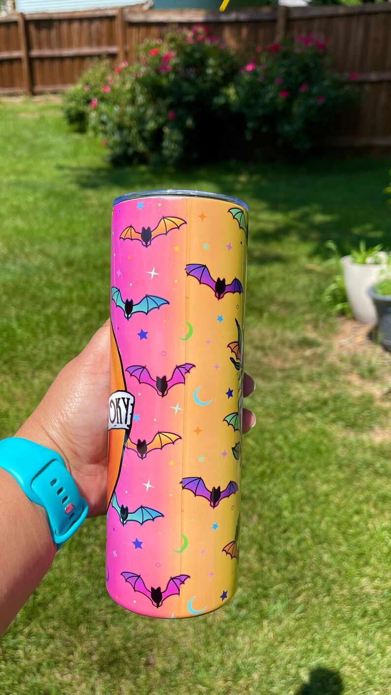 May include: A Halloween-themed tumbler with a gradient background of pink, orange, and yellow. The tumbler is decorated with colorful bats, stars, and crescent moons. The word "Spooky" is printed on the tumbler.