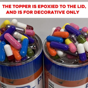 May include: Two clear glass cups filled with colorful capsules. The cups are blue and red with white text. The text on the cups reads "THE TOPPER IS EPOXIED TO THE LID, AND IS FOR DECORATIVE ONLY".