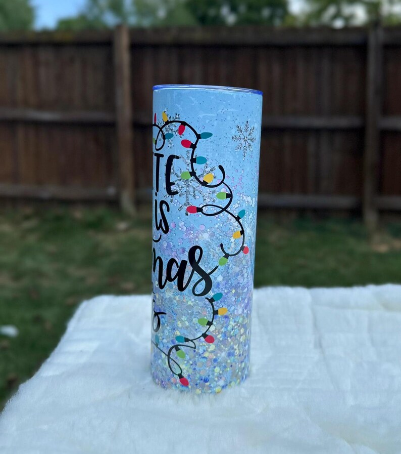 May include: A blue and white tumbler with a glitter finish. The tumbler has a design of colorful Christmas lights and the text "It is Christmas".