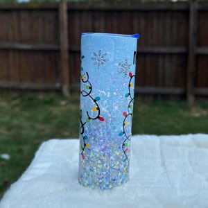 May include: A blue and white tumbler with a snowflake design and colorful Christmas lights. The tumbler is covered in glitter.