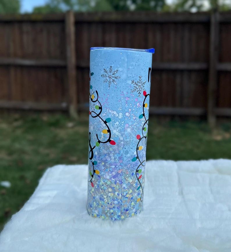 May include: A blue and white tumbler with a snowflake design and colorful Christmas lights. The tumbler is covered in glitter.