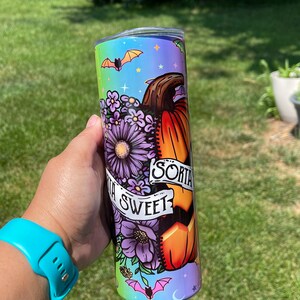 May include: A colorful tumbler with a Halloween theme. The design features a pumpkin, purple flowers, and bats against a rainbow background. The text "Sorta Sweet" is displayed on a banner. The tumbler is held by a person wearing a blue watch.