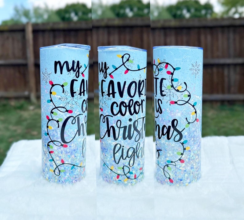 May include: A blue tumbler with glitter and a white snowflake design. The tumbler has the text "My favorite color is Christmas lights" in black.