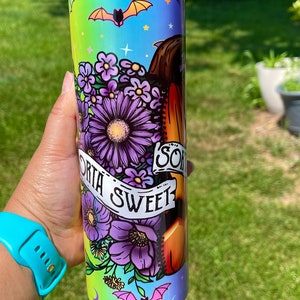 May include: A tall, insulated tumbler with a colorful design featuring purple flowers, bats, and the words "Sorta Sweet." The tumbler has a gradient background of green, blue, and purple. The design wraps around the entire tumbler.