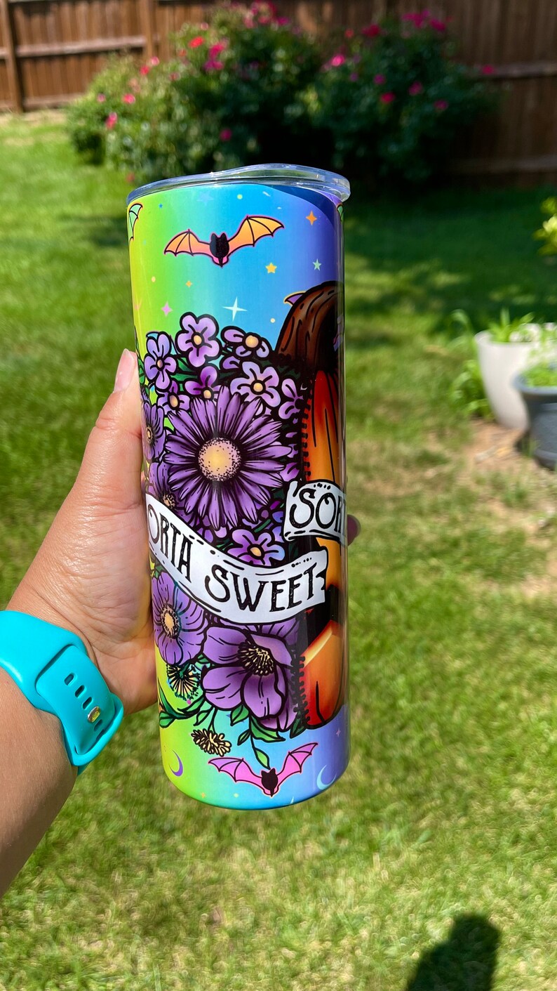 May include: A tall, insulated tumbler with a colorful design featuring purple flowers, bats, and the words "Sorta Sweet." The tumbler has a gradient background of green, blue, and purple. The design wraps around the entire tumbler.