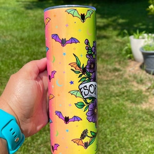 May include: A tall, cylindrical tumbler with a colorful design featuring bats, stars, and moons on a gradient yellow to pink background. The tumbler has a floral design with the word "SOR" on a banner. The tumbler is held by a person.