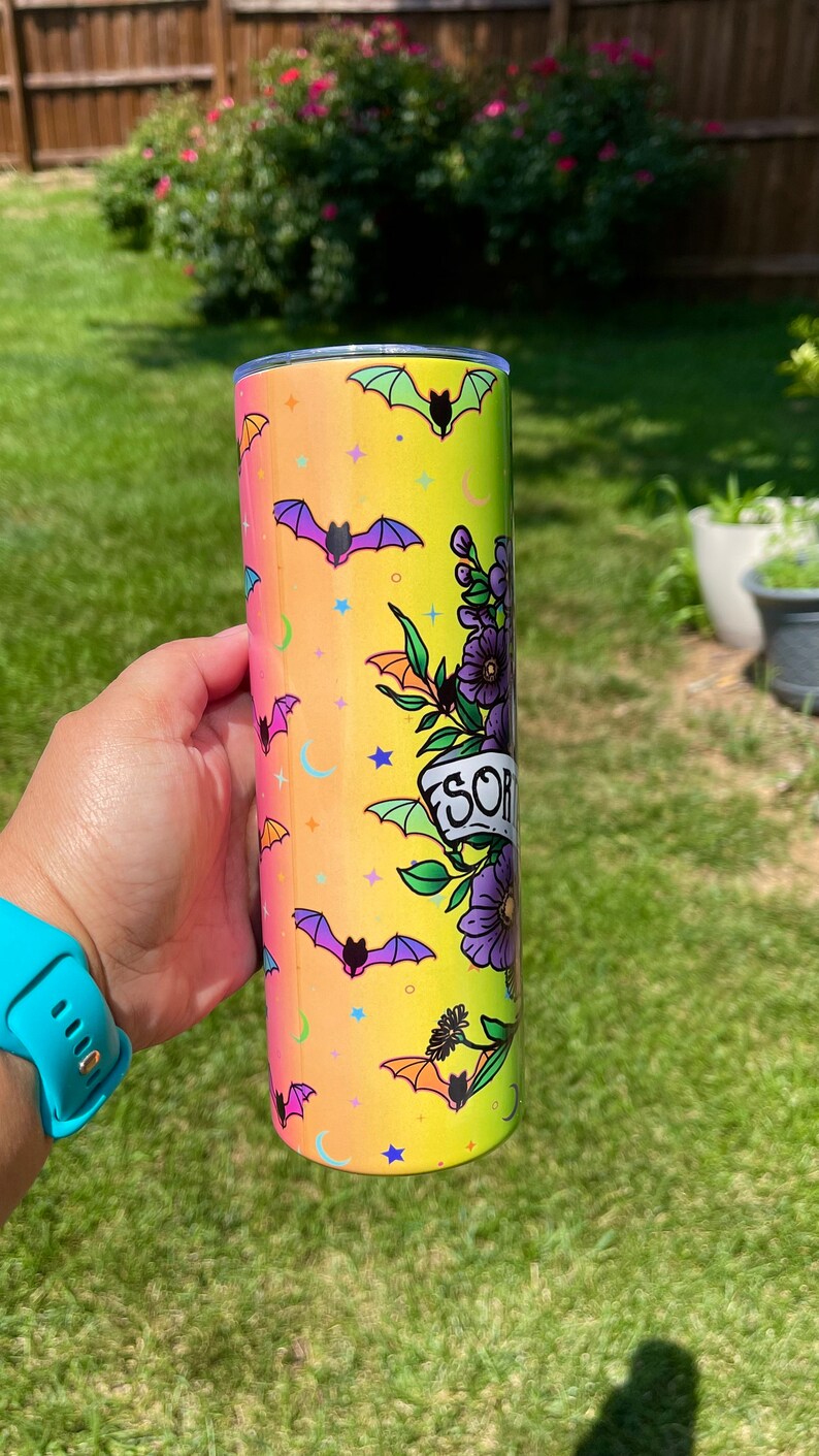 May include: A tall, cylindrical tumbler with a colorful design featuring bats, stars, and moons on a gradient yellow to pink background. The tumbler has a floral design with the word "SOR" on a banner. The tumbler is held by a person.