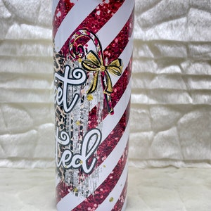 May include: A stainless steel tumbler with a candy cane design. The tumbler features red glitter stripes and white stripes. The text "Sweet Limited" is written on the tumbler, along with a candy cane and a gold bow.