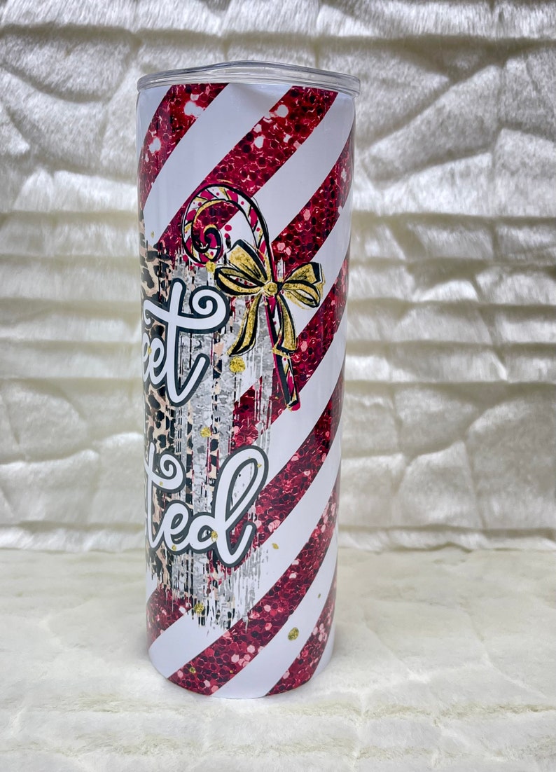 May include: A stainless steel tumbler with a candy cane design. The tumbler features red glitter stripes and white stripes. The text "Sweet Limited" is written on the tumbler, along with a candy cane and a gold bow.