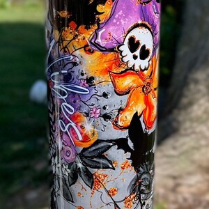 May include: A tall, cylindrical tumbler with a Halloween-themed design. The design features a skull with heart-shaped eyes, bats, flowers, and spiderwebs in shades of purple, orange, black, and gray. The word "Spooky" is written on the tumbler.