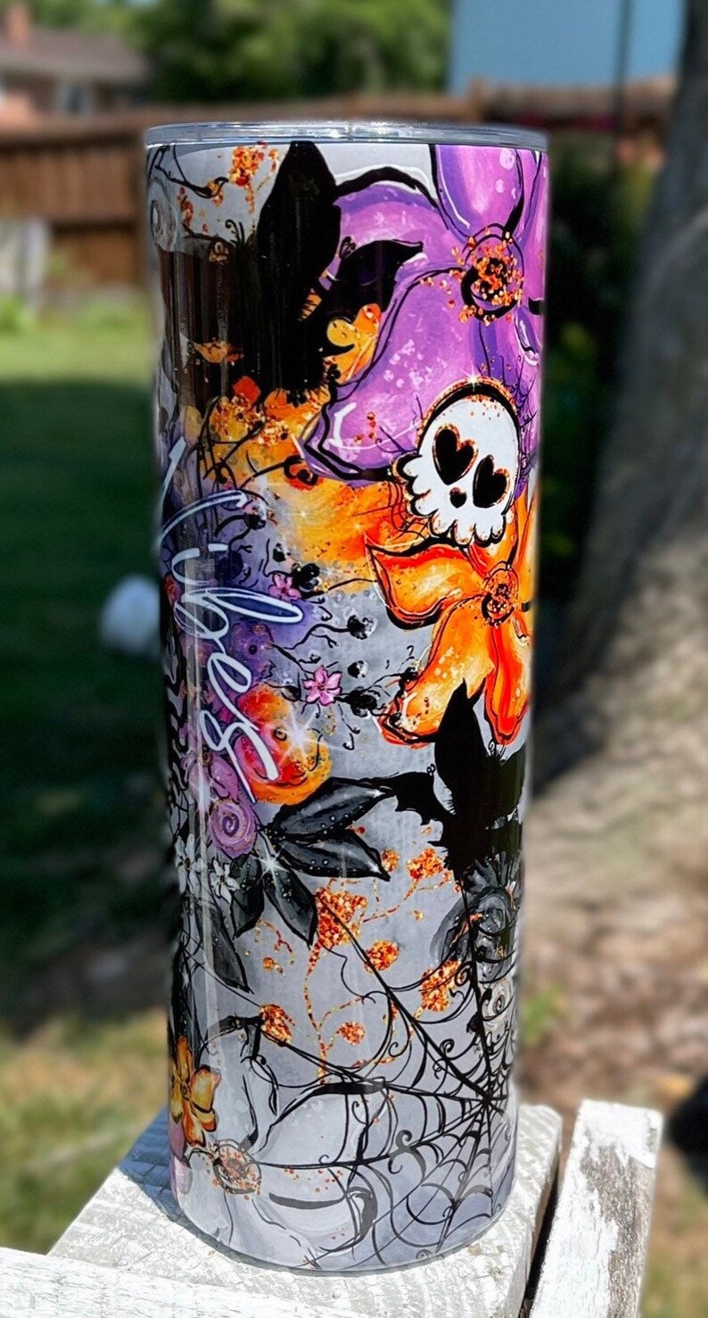 May include: A tall, cylindrical tumbler with a Halloween-themed design. The design features a skull with heart-shaped eyes, bats, flowers, and spiderwebs in shades of purple, orange, black, and gray. The word "Spooky" is written on the tumbler.