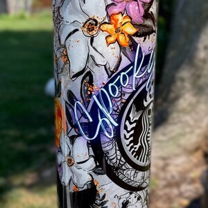 May include: A tall, insulated tumbler with a spooky floral design. The tumbler features white flowers, purple and orange accents, spiderwebs, and the word "Spooky" in a cursive font. A skull and Starbucks logo are also visible.