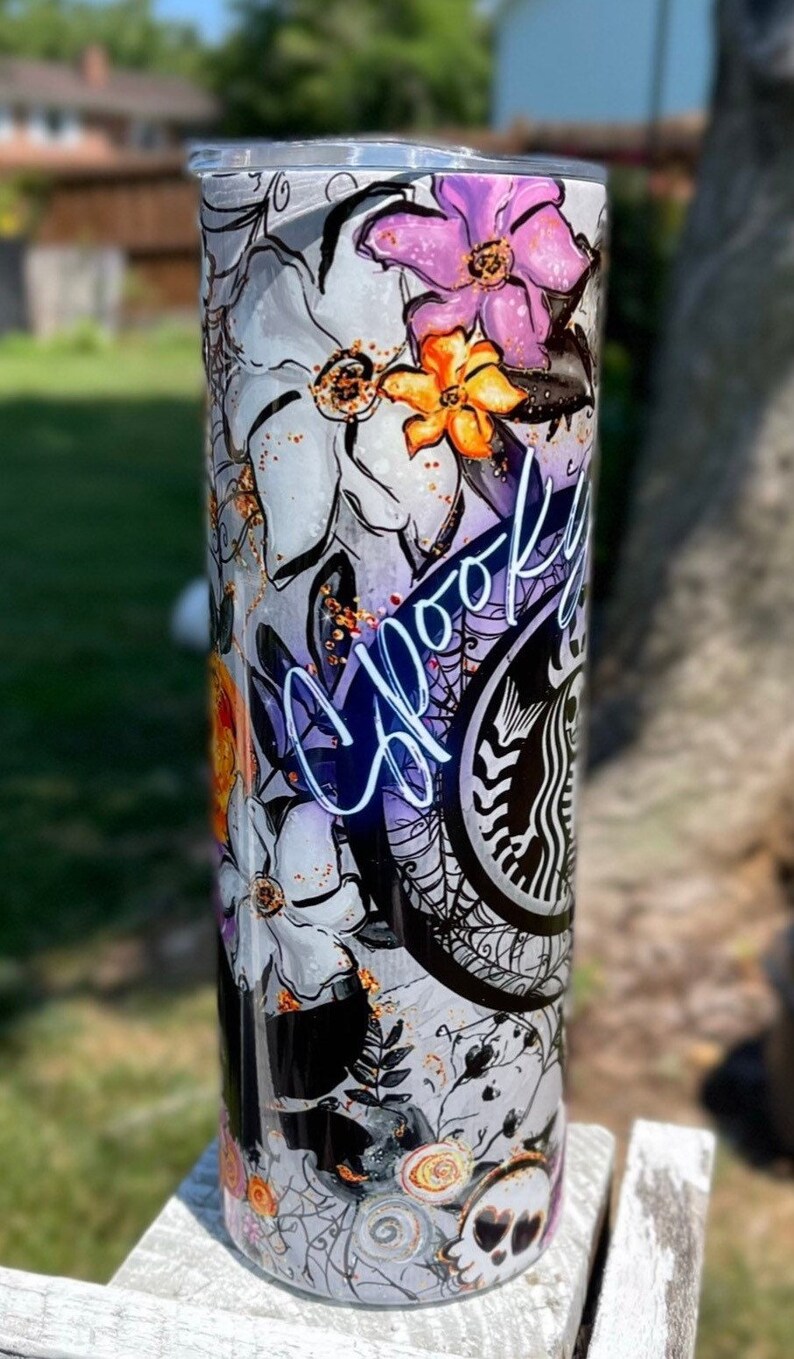 May include: A tall, insulated tumbler with a spooky floral design. The tumbler features white flowers, purple and orange accents, spiderwebs, and the word "Spooky" in a cursive font. A skull and Starbucks logo are also visible.