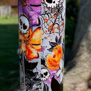 May include: A tall, cylindrical tumbler with a Halloween-themed design. The tumbler features a repeating pattern of skulls, spiderwebs, orange and purple flowers, and black bats. The background is a light gray color.