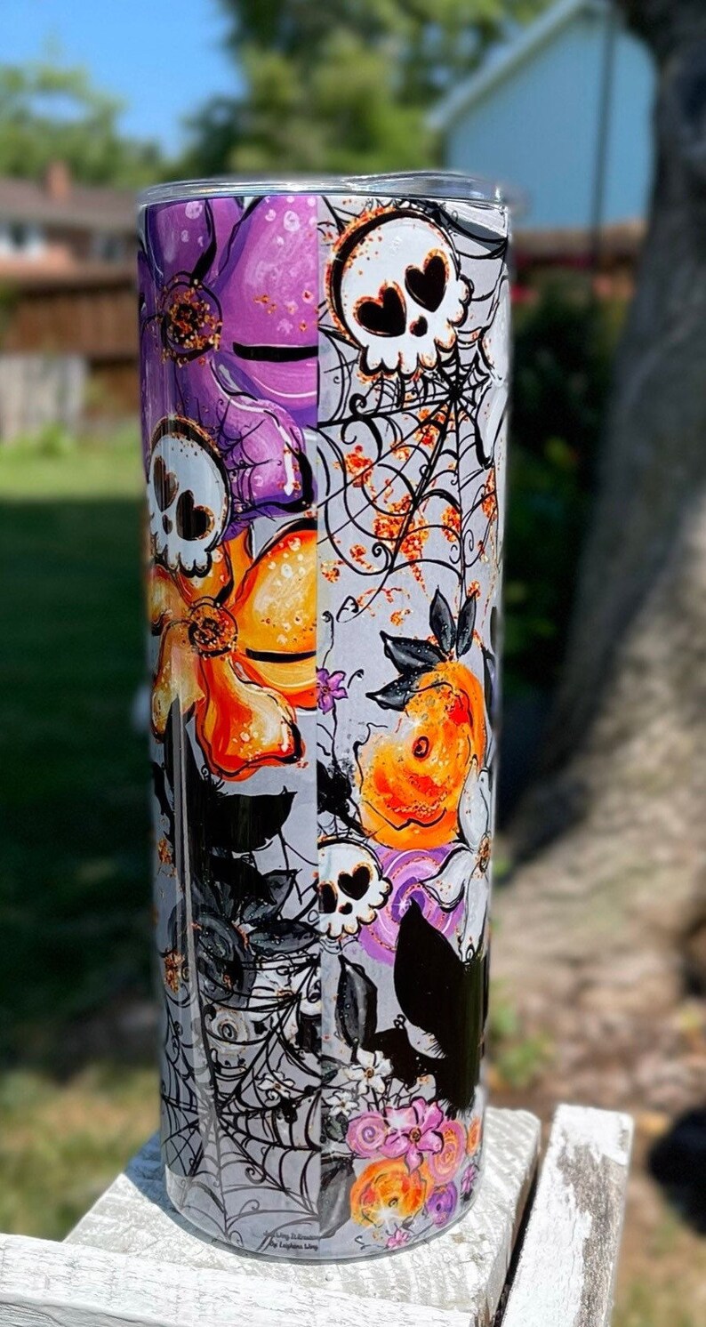 May include: A tall, cylindrical tumbler with a Halloween-themed design. The tumbler features a repeating pattern of skulls, spiderwebs, orange and purple flowers, and black bats. The background is a light gray color.